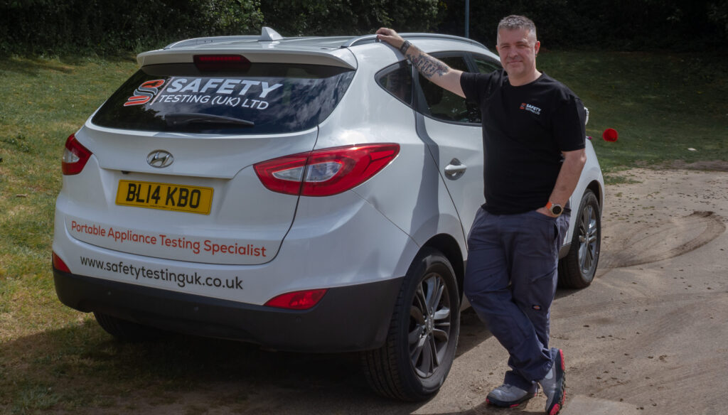 A technician in black shirt and blue trousers leans on a white van displaying Safety Testing UK branding, pat testing and electronics testing services, smiling in an outdoor setting