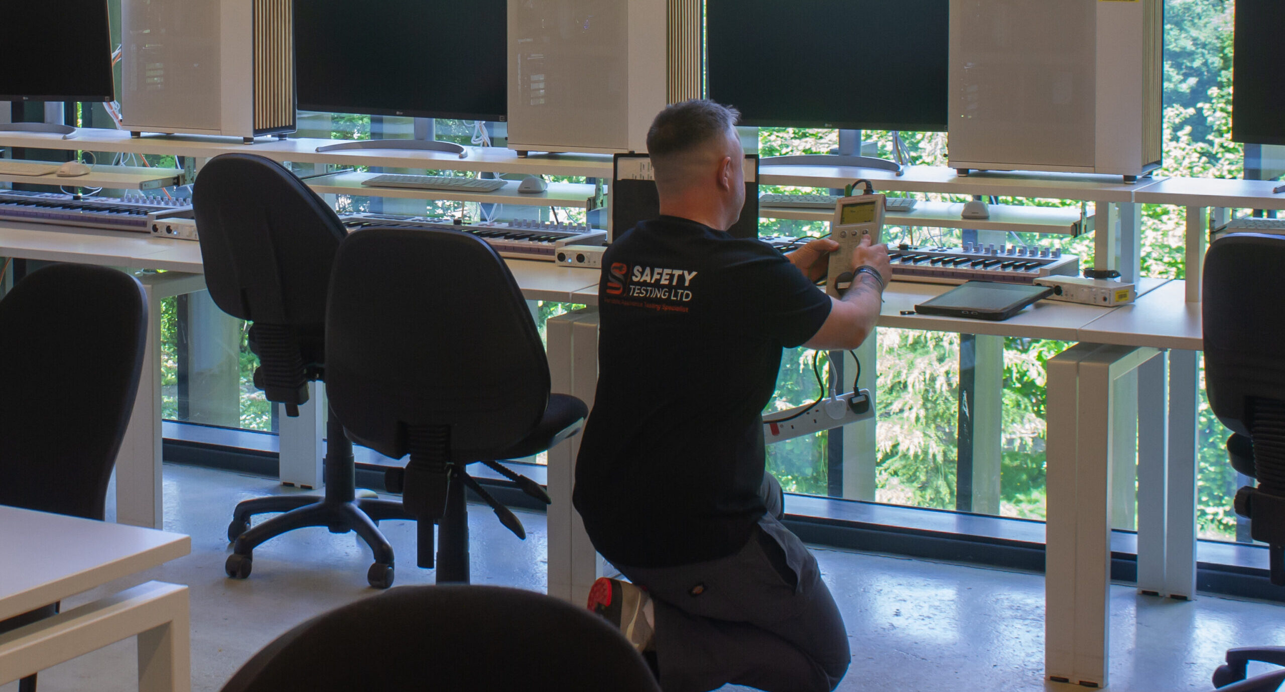 A technician in a black shirt kneels beside white desks, performing pat testing on electronics in a bright office with large windows and green views outside.