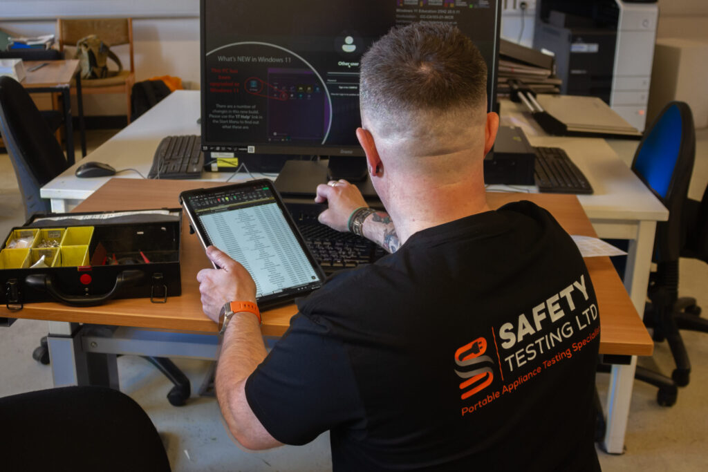A technician in a black Safety Testing Ltd shirt performs electronics testing on a tablet at a desk