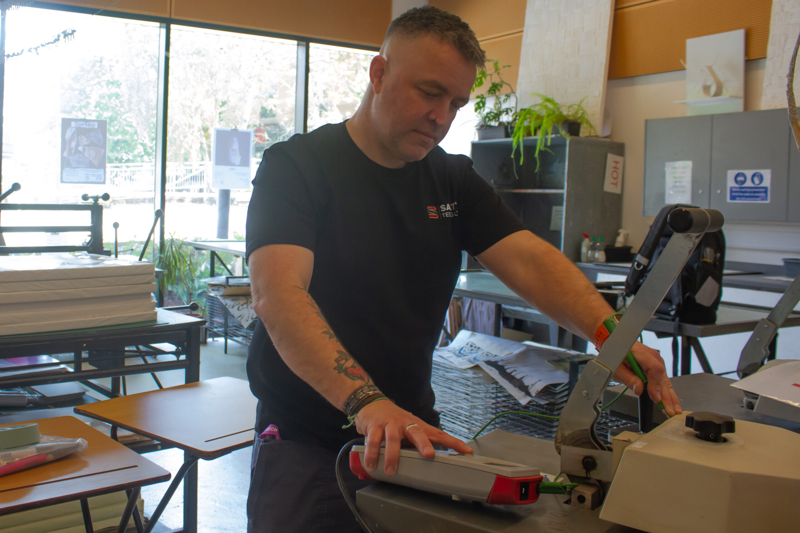 A man in a black shirt performs pat testing on an electrical device in a workshop setting with tools and equipment around
