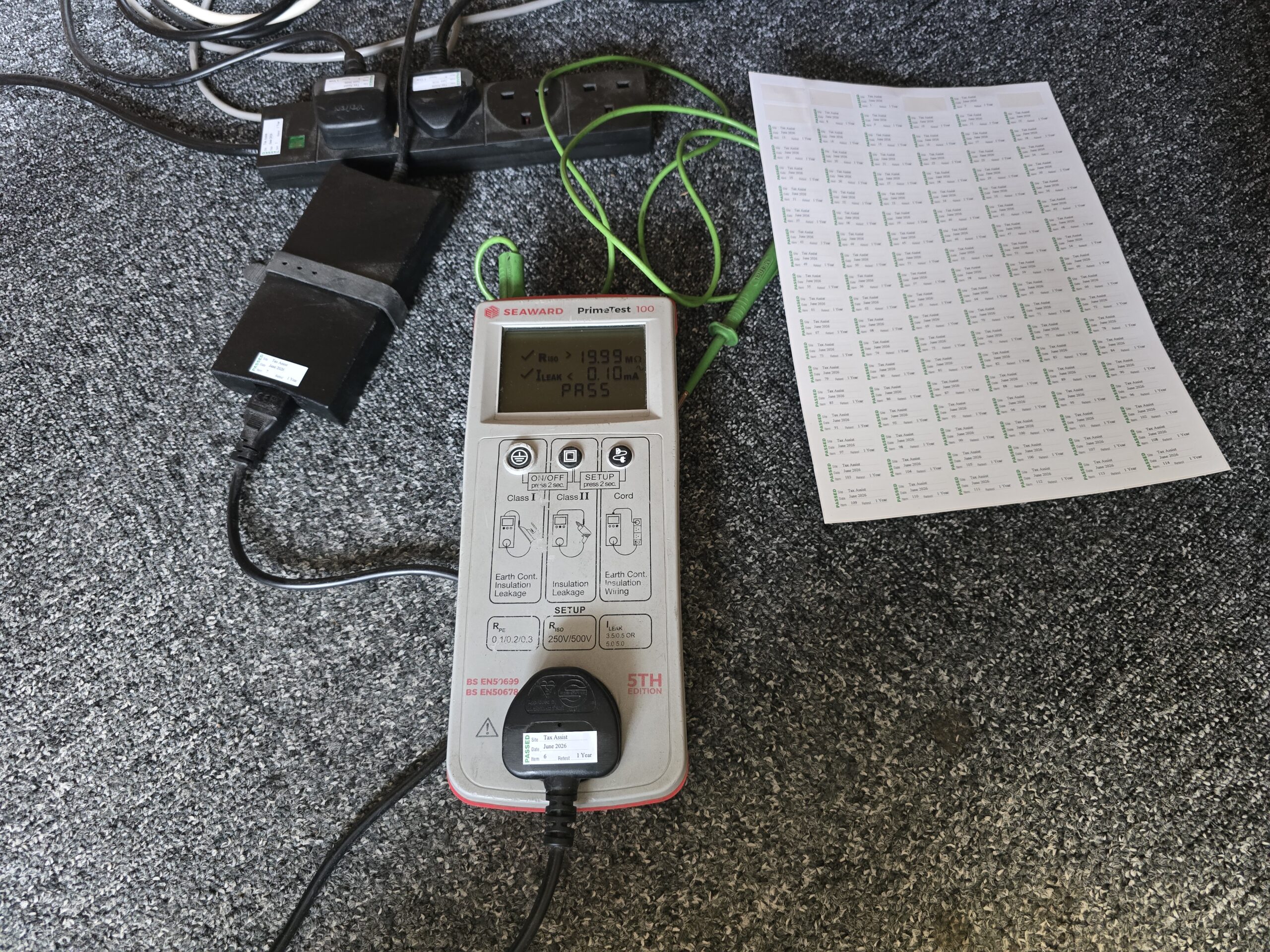 A Seaward PrimeTest 100 device performs pat testing on black power strips and cables on grey carpet.