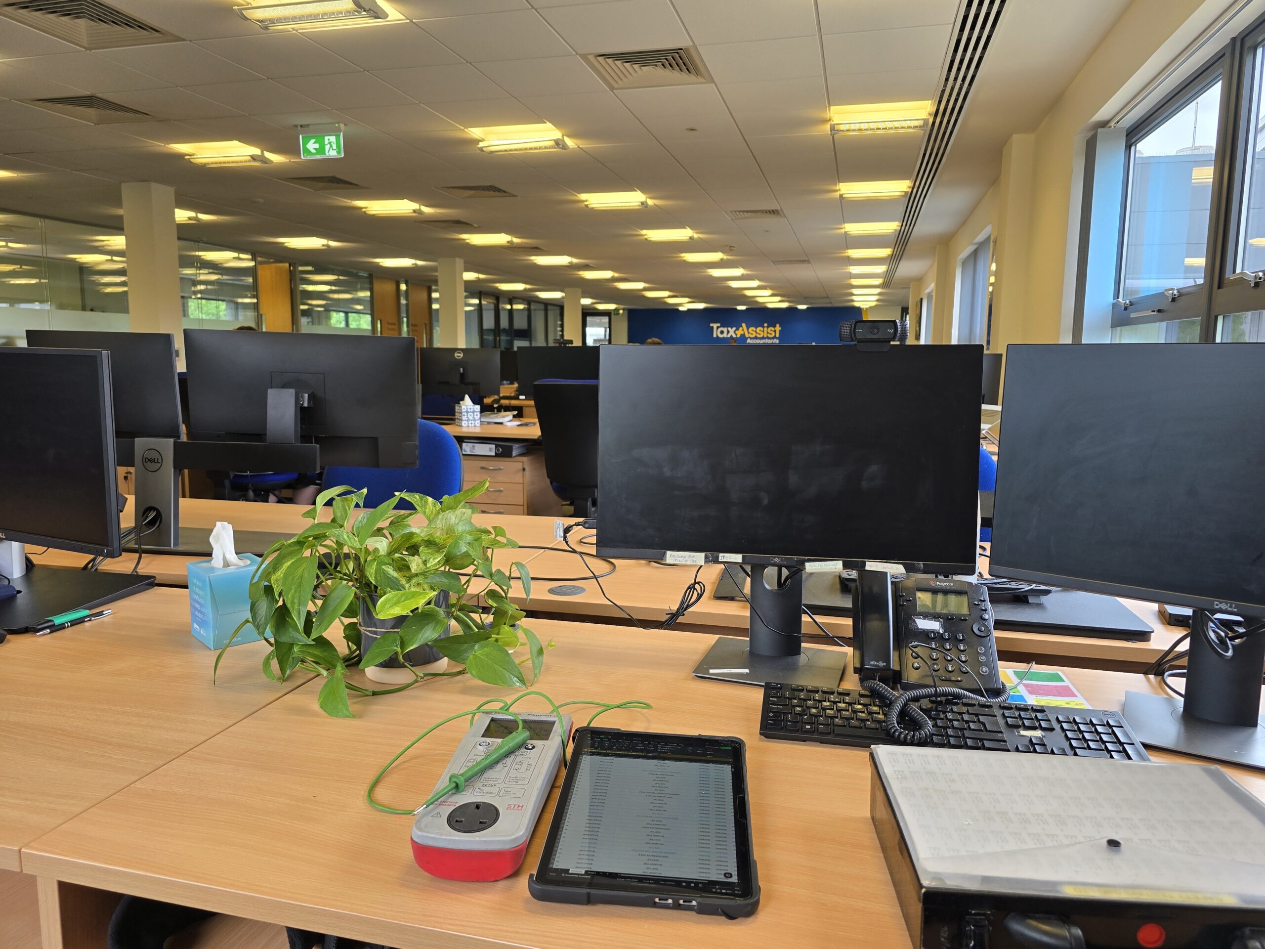 A bright office scene featuring a wooden desk with computer monitors, a potted plant, and tools for pat testing and electronics testing.